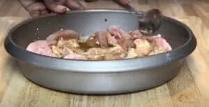 Person marinating raw chicken pieces in a pan on a wooden surface.